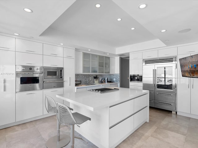 kitchen with built in appliances, a kitchen bar, a raised ceiling, and white cabinetry