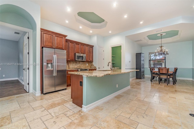 kitchen featuring appliances with stainless steel finishes, light stone counters, a tray ceiling, an island with sink, and decorative backsplash