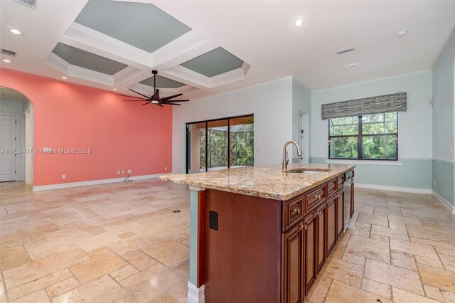 kitchen with sink, coffered ceiling, a kitchen island with sink, ceiling fan, and light stone countertops