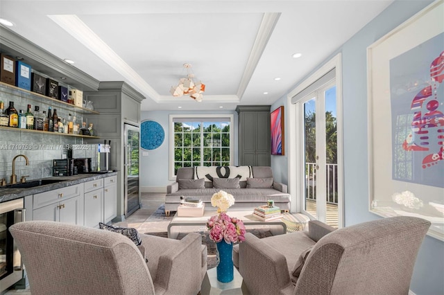 living room featuring a raised ceiling, a notable chandelier, beverage cooler, wet bar, and ornamental molding