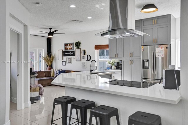 kitchen with stainless steel refrigerator with ice dispenser, gray cabinets, kitchen peninsula, island range hood, and a breakfast bar