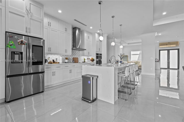 kitchen with wall chimney exhaust hood, white cabinetry, stainless steel appliances, hanging light fixtures, and a kitchen island with sink