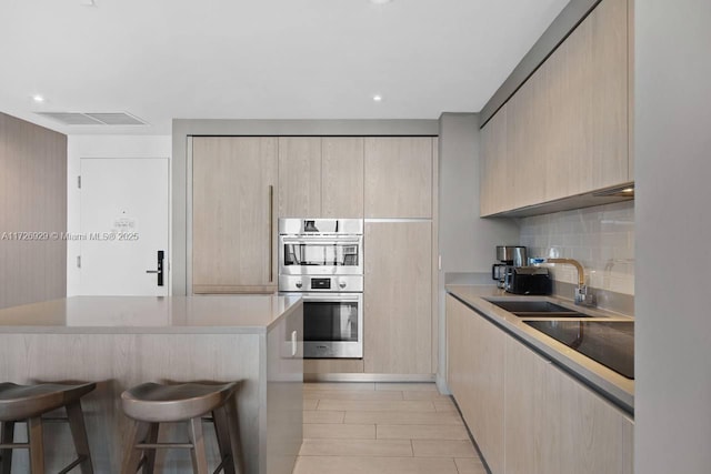 kitchen with light brown cabinets, double oven, decorative backsplash, a kitchen breakfast bar, and sink