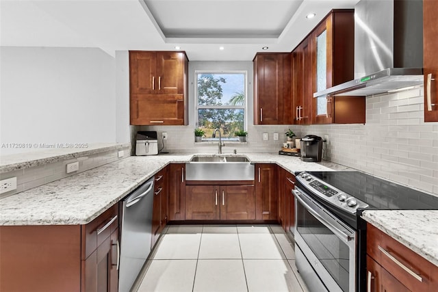 kitchen featuring stainless steel appliances, a tray ceiling, wall chimney exhaust hood, light stone counters, and sink