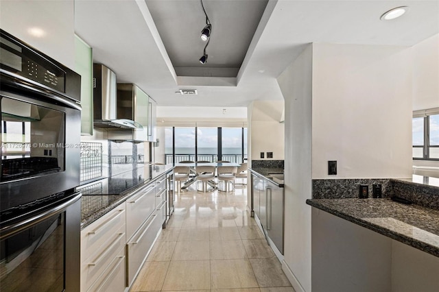 kitchen featuring white cabinets, track lighting, wall chimney exhaust hood, dark stone counters, and a raised ceiling