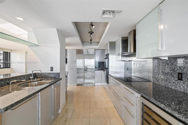 kitchen with rail lighting, stainless steel built in fridge, dark stone countertops, wall chimney range hood, and white cabinets