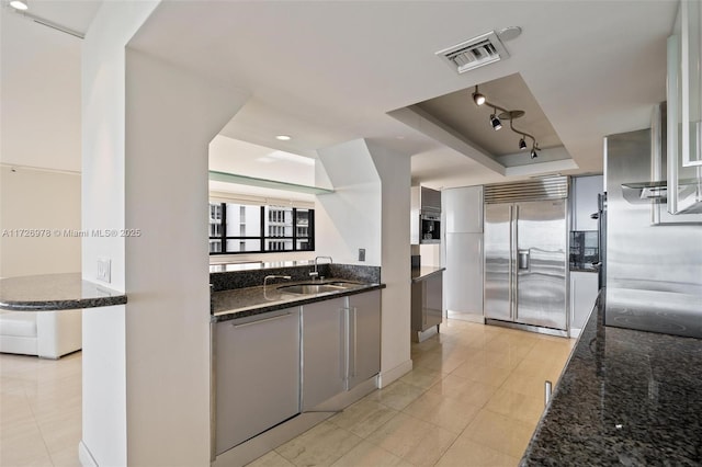 kitchen featuring dishwasher, sink, a tray ceiling, built in refrigerator, and dark stone counters