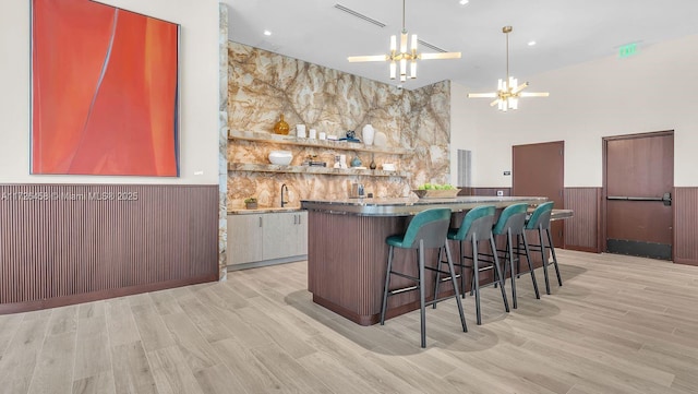 bar featuring light wood-style flooring, a wainscoted wall, a towering ceiling, hanging light fixtures, and an inviting chandelier