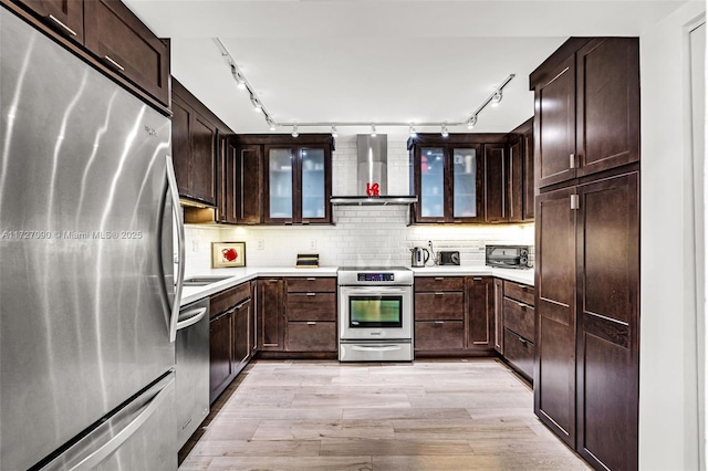 kitchen with dark brown cabinetry, appliances with stainless steel finishes, wall chimney exhaust hood, decorative backsplash, and light hardwood / wood-style flooring