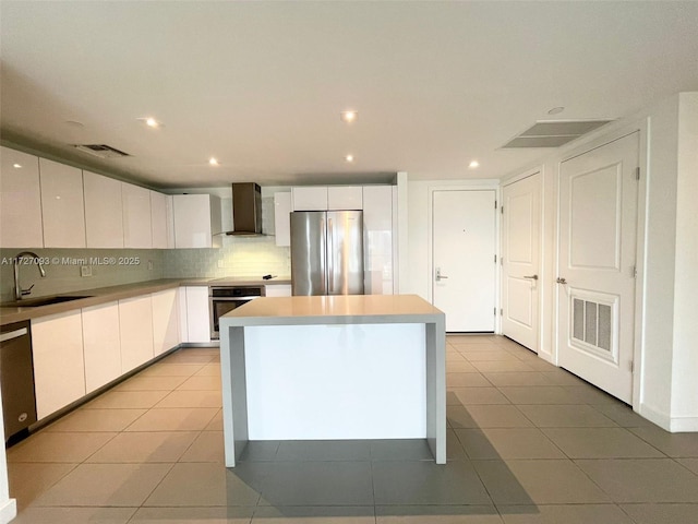 kitchen with wall chimney range hood, a kitchen island, sink, white cabinetry, and stainless steel appliances