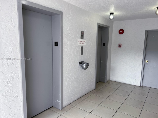 corridor with light tile patterned floors, a textured wall, a textured ceiling, and elevator