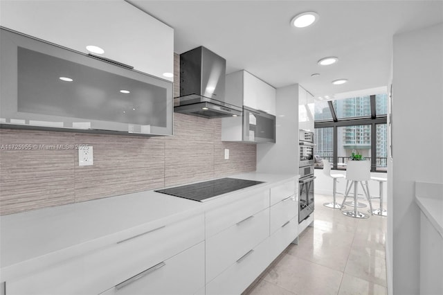 kitchen featuring wall chimney range hood, black electric stovetop, decorative backsplash, light tile patterned floors, and white cabinets