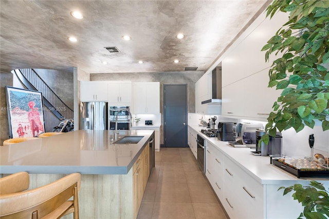 kitchen with a large island, wall chimney range hood, sink, white cabinetry, and stainless steel appliances