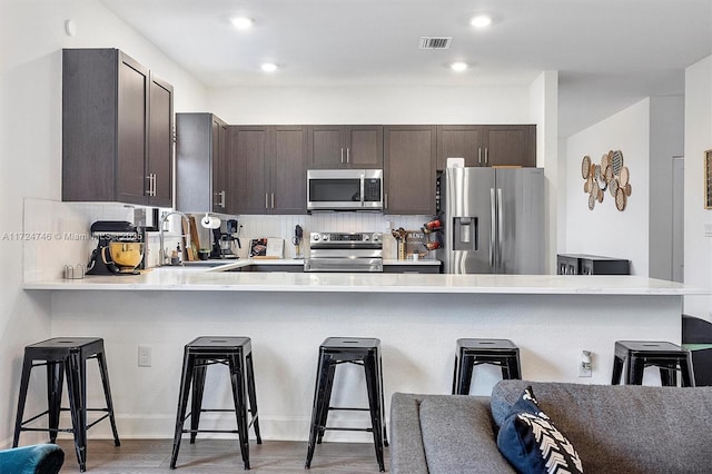 kitchen with dark brown cabinetry, appliances with stainless steel finishes, a kitchen bar, kitchen peninsula, and hardwood / wood-style flooring