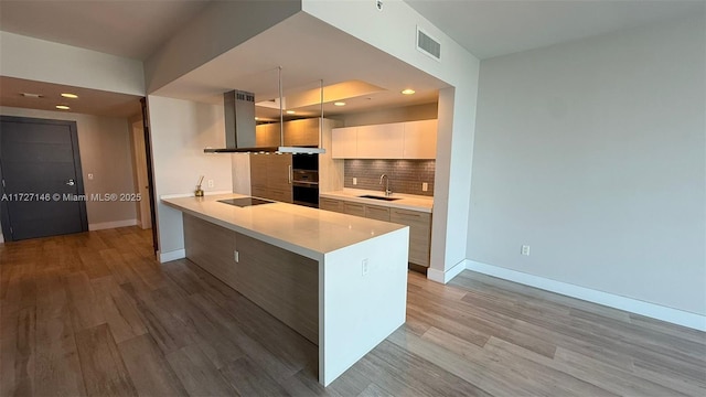 kitchen featuring island range hood, backsplash, black electric cooktop, light wood-type flooring, and sink