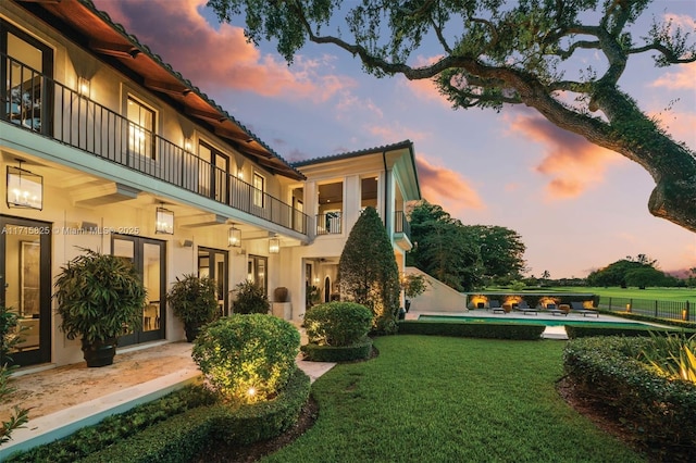 back house at dusk featuring french doors, a balcony, a swimming pool, and a lawn