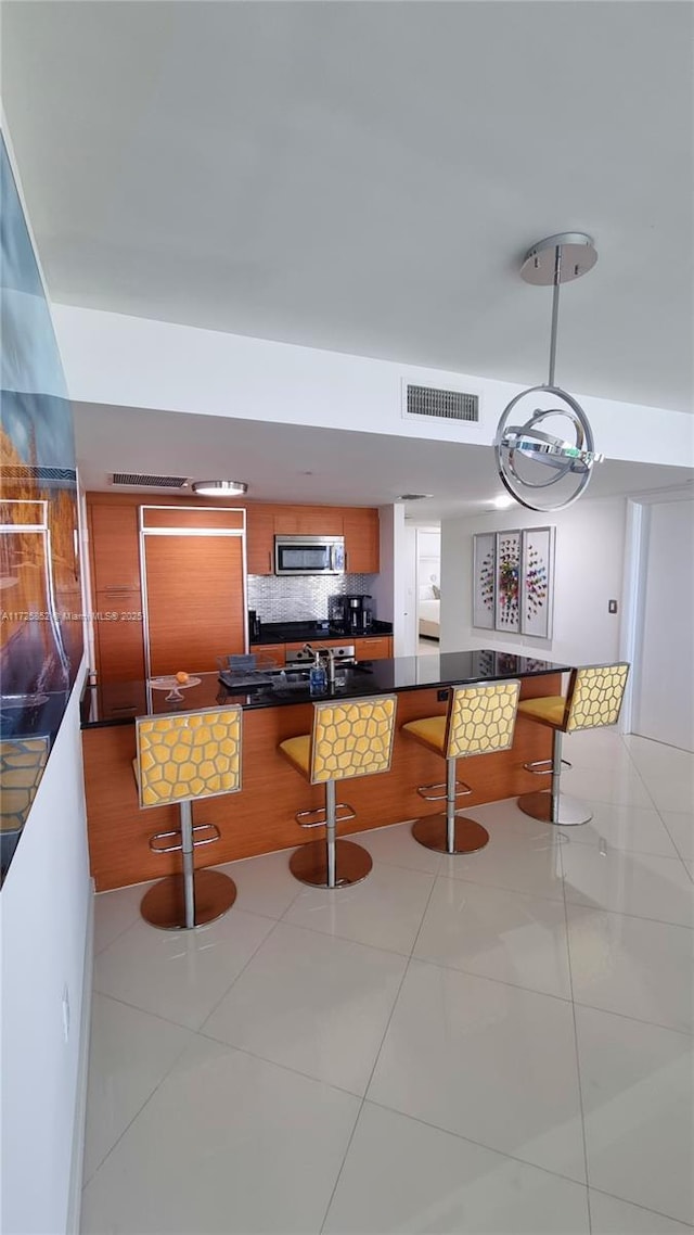 50 Biscayne Blvd Unit 1902, Miami, FL 33132 - kitchen featuring stainless steel microwave, a breakfast bar area, dark countertops, brown cabinetry, and light tile patterned floors