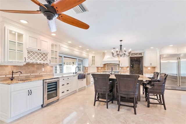 kitchen featuring wine cooler, pendant lighting, a center island with sink, built in refrigerator, and ceiling fan with notable chandelier