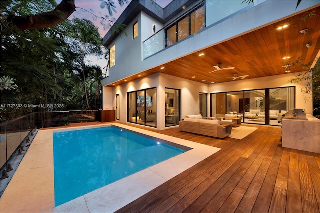 pool at dusk featuring exterior kitchen, a wooden deck, an outdoor living space, and ceiling fan