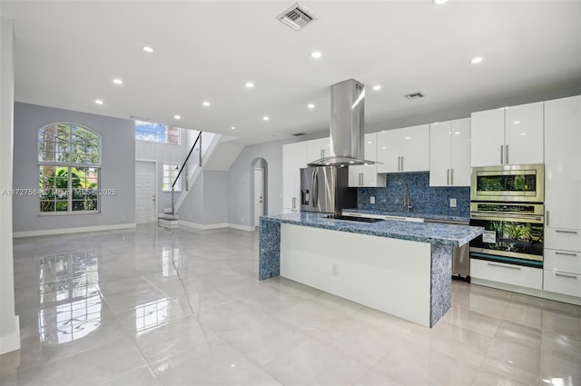 kitchen with a center island with sink, appliances with stainless steel finishes, white cabinets, dark stone counters, and island range hood