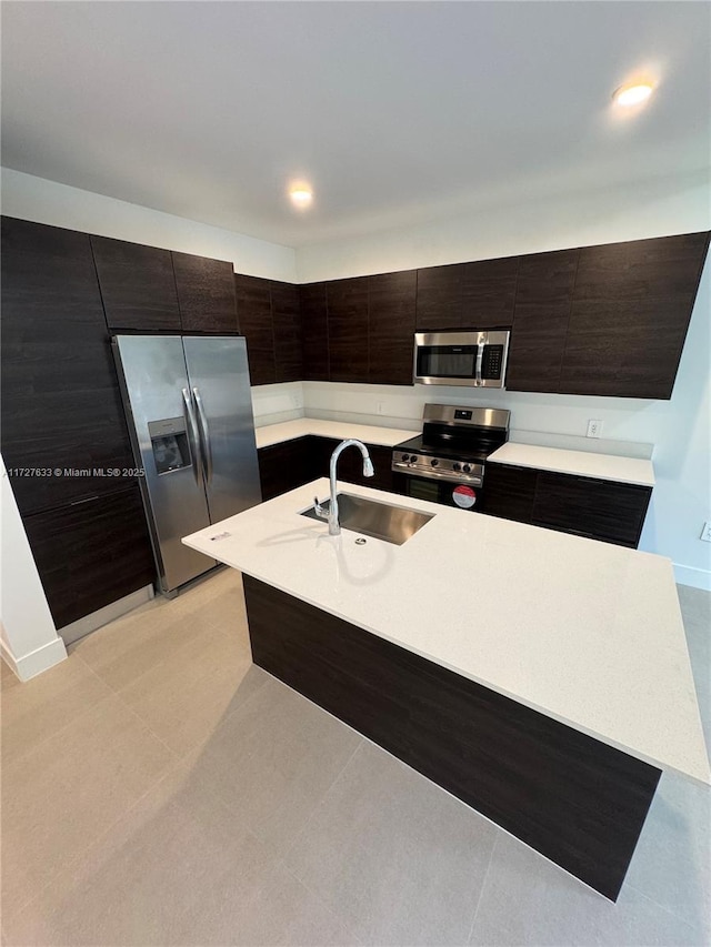 kitchen with light tile patterned floors, appliances with stainless steel finishes, a kitchen island with sink, dark brown cabinets, and sink