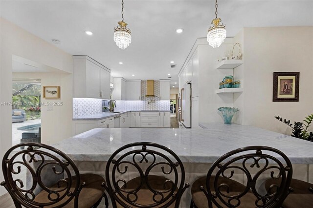 kitchen featuring hanging light fixtures, wall chimney range hood, white cabinets, and decorative backsplash