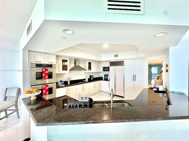 kitchen with double oven, sink, paneled built in fridge, a raised ceiling, and wall chimney range hood