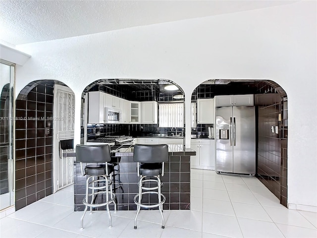 kitchen featuring light tile patterned floors, a kitchen bar, stainless steel fridge, a textured ceiling, and white cabinets