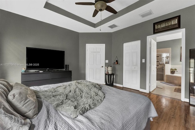 bedroom with a raised ceiling, wood-type flooring, connected bathroom, and ceiling fan