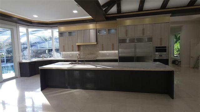 kitchen featuring crown molding, backsplash, stainless steel appliances, an island with sink, and light brown cabinetry