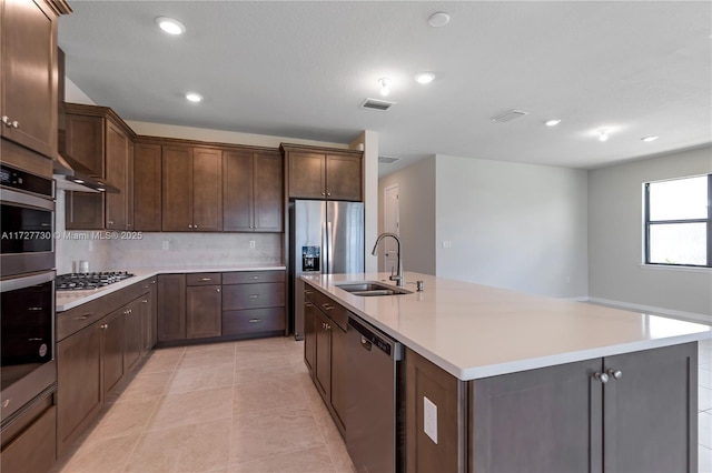 kitchen with dark brown cabinetry, stainless steel appliances, sink, light tile patterned floors, and a center island with sink