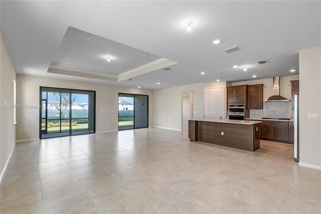 kitchen featuring a raised ceiling, decorative backsplash, gas cooktop, a kitchen island with sink, and wall chimney exhaust hood