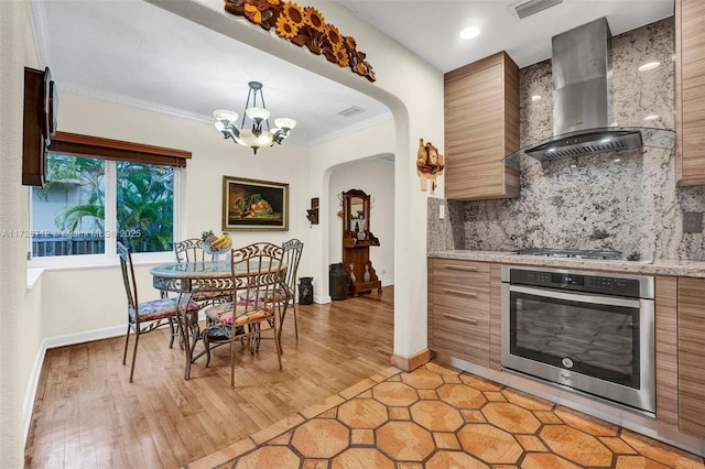 kitchen with crown molding, appliances with stainless steel finishes, tasteful backsplash, decorative light fixtures, and wall chimney exhaust hood