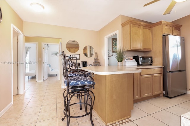 kitchen with light tile patterned floors, kitchen peninsula, ceiling fan, a kitchen breakfast bar, and stainless steel refrigerator