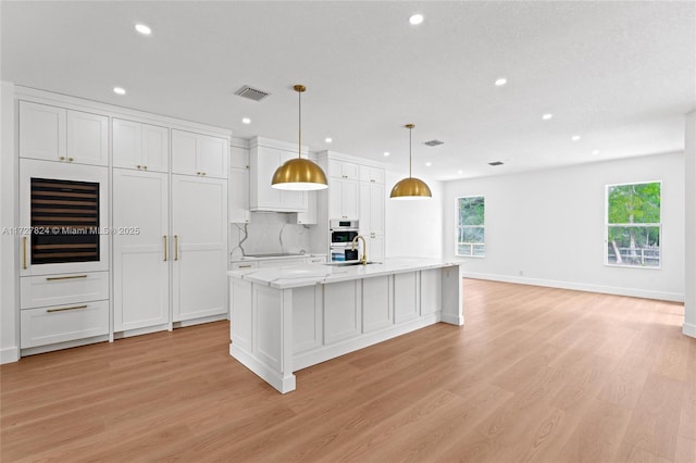 kitchen with a kitchen island with sink, light wood-type flooring, hanging light fixtures, white cabinets, and sink