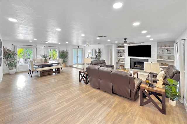 living room with ceiling fan, built in shelves, light wood-type flooring, and a large fireplace