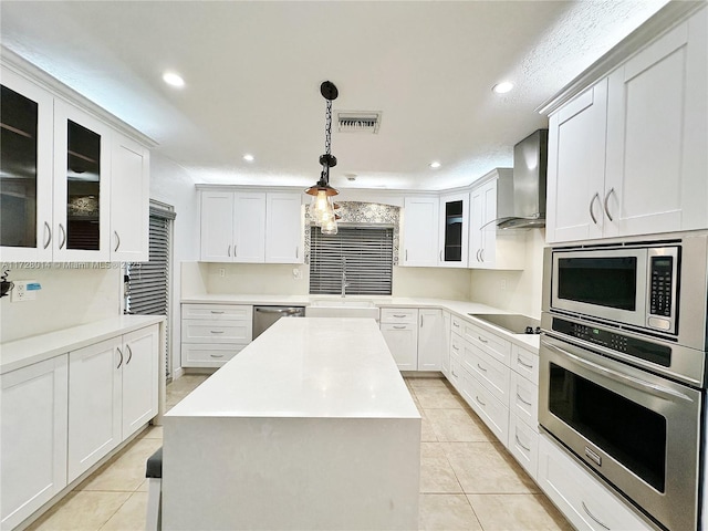 kitchen featuring white cabinetry, appliances with stainless steel finishes, decorative light fixtures, wall chimney range hood, and a center island