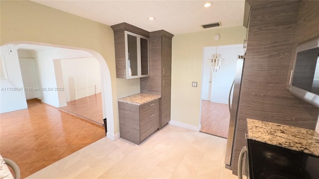 kitchen with a notable chandelier, appliances with stainless steel finishes, light tile patterned floors, dark brown cabinets, and light stone counters