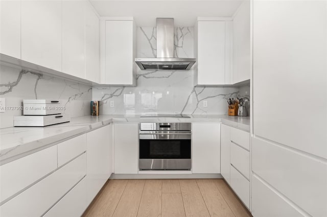 kitchen with white cabinets, oven, light wood-type flooring, and wall chimney range hood