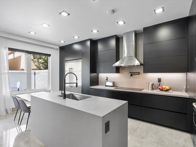 kitchen with wall chimney exhaust hood, black electric stovetop, sink, backsplash, and a kitchen island with sink