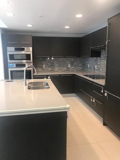 kitchen featuring light tile patterned floors, stainless steel double oven, an island with sink, tasteful backsplash, and sink