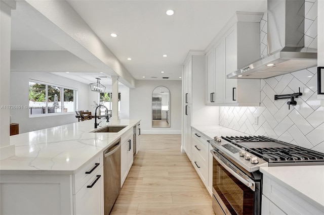 kitchen with sink, white cabinetry, a kitchen island with sink, stainless steel appliances, and wall chimney exhaust hood