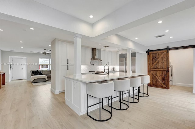 kitchen with a barn door, a kitchen bar, wall chimney range hood, a large island with sink, and white cabinets