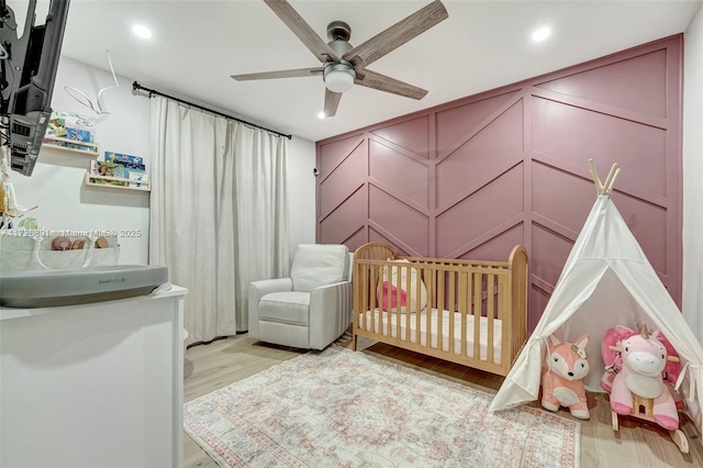 bedroom with light wood-type flooring, ceiling fan, and a nursery area