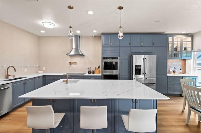 kitchen featuring sink, a kitchen island with sink, appliances with stainless steel finishes, wall chimney exhaust hood, and light stone counters