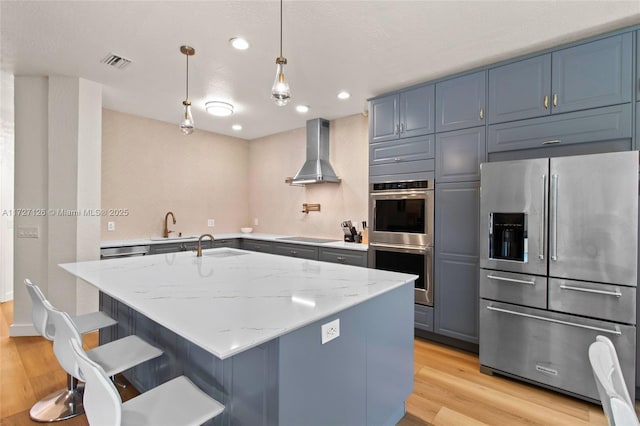 kitchen featuring stainless steel appliances, a kitchen island with sink, decorative light fixtures, light stone countertops, and wall chimney range hood