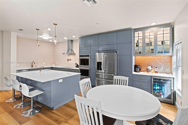 kitchen with appliances with stainless steel finishes, beverage cooler, hanging light fixtures, wall chimney exhaust hood, and light hardwood / wood-style flooring