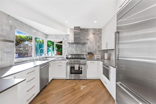 kitchen with premium appliances, light hardwood / wood-style floors, wall chimney range hood, white cabinets, and sink