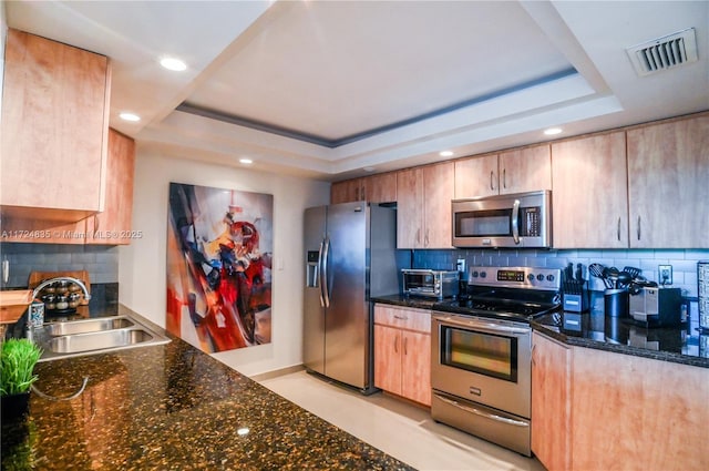 kitchen featuring backsplash, dark stone countertops, a raised ceiling, sink, and stainless steel appliances