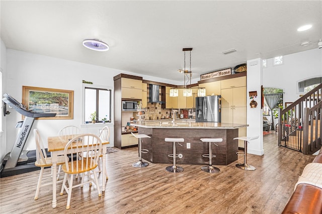 kitchen featuring wall chimney range hood, a kitchen bar, hanging light fixtures, light wood-type flooring, and appliances with stainless steel finishes